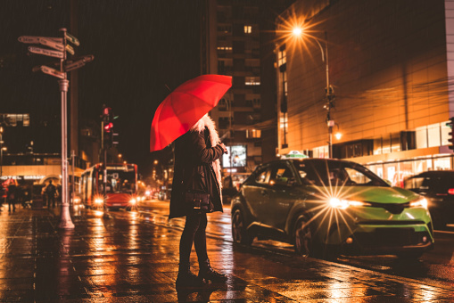 Woman with red umbrella standing downtown at night waiting green light at crosswalk.