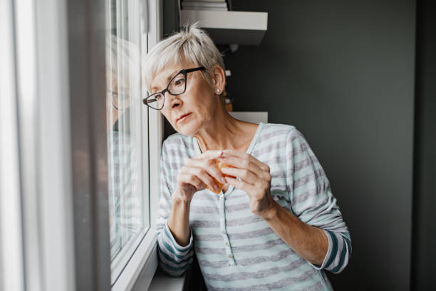 istock Sad senior woman at home, looking through the window 1356372505