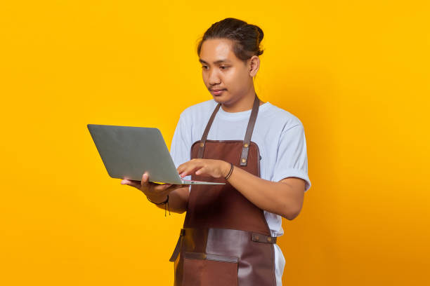 istock Portrait of handsome young barista using laptop looking serious doing office work on yellow background 1356374075