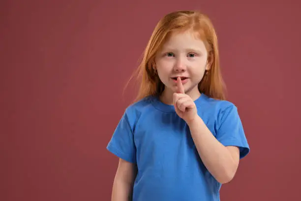 Redhead girl in blue t-shirt putting finger up to lips saying shahs on red background