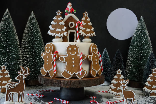 Stock photo showing close-up view of a night-time Christmas forest scene designed Christmas cake. Homemade, cake covered in fondant icing and candy cane holding gingerbread men, decorated with white royal icing, topped by a gingerbread house with Father Christmas on the roof. The cake is displayed on a wooden cake stand surrounded by model fir trees, reindeer and Christmas tree shaped cookies, against a black background with a full moon. imagen casera, exhibición de pasteles navideños, pastel cubierto con glaseado de fondant, cubierto con galletas de la casa de pan de jengibre heladas con glaseado real blanco, hombres de pan de jengibre y bastones de caramelo, escena nocturna del bosque