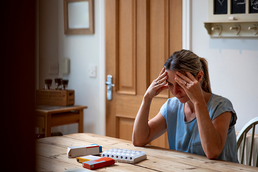 A mid adult woman sitting at her dining table at home, organising her daily medication. She is looking at the pill boxes on the table while she has her head in her hands, worried about her medication.