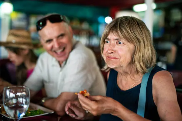 Senior woman tries a new dish in a restaurant. She is finding the new taste disgusting. Senior man is laughing at her.