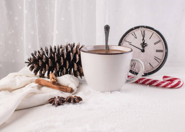 시계, 전나무 콘, 커피 머그잔, 스타 아니스, 계피 스틱 및 롤리팝이있는 흰색 눈 테이블. 새해 개념. 시계의 손은 거의 12시를 보여줍니다. - 12 oclock 뉴스 사진 이미지
