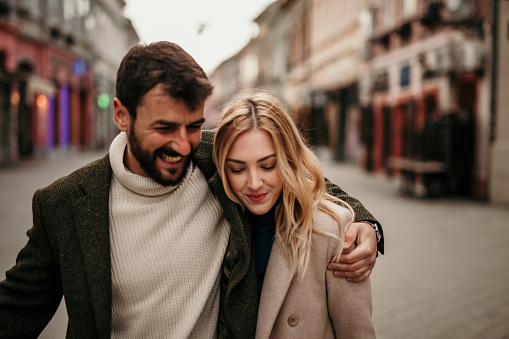 Embraced stylishly dressed couple walking through the city, both wearing coats and having a great time.