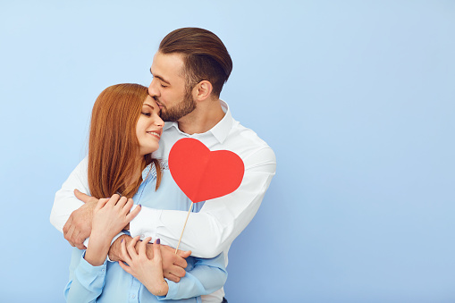 Side view of attractive young man and woman with adorable paper hearts smiling and looking at camera while standing on blue background