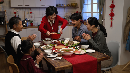cheerful asian senior woman grandmother in red clothing putting thumb up while serving her specialty on table, feeling proud of her cooking. ready to have family reunion dinner on lunar new year