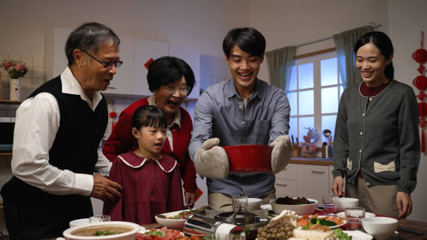 smiling asian father serving hot soup on the dining table and posing with folded arms, feeling proud of his cooking. preparing food for family on lunar new year’s eve