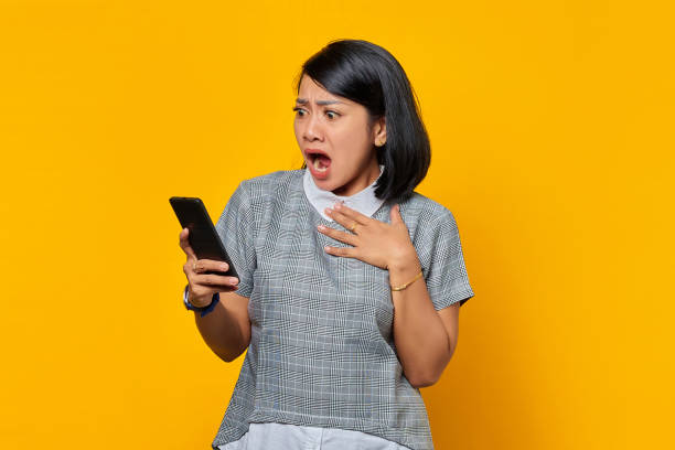 jeune femme asiatique surprise tenant un téléphone portable la bouche ouverte sur fond jaune - mouth open women displeased anger photos et images de collection