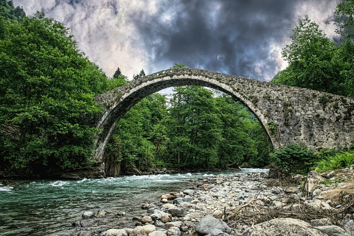 Picturesque countryside in the Balkans. Ancient medieval arched stone bridge and mountain river against the backdrop of green mountain slopes and purple sunset sky