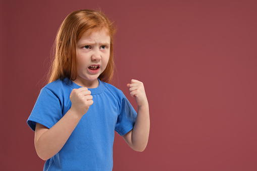 close-up portrait of cute redhead emotional little girl in blue t-shirt on red background. She is about to get nervous, angry with someone