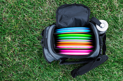 Bag of multi-coloured flying discs used for disc golf on green grass.