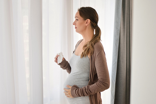 Portrait of a worried young pregnant woman standing by the window, holding her belly