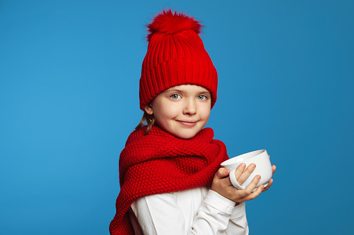 Adorable child wearing knitted red scarf and hat warming hands while holding cup of tea, isolated on blue background