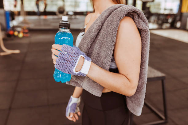 Young unrecognizable fitness woman drinking isotonic drink Young attractive unrecognizable fitness woman with isotonic drink in her hand at gym after exercise. Physical exercise and healthy lifestyle concept. sport drink stock pictures, royalty-free photos & images