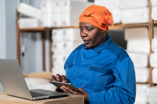 istock Female warehouse worker   checking information on  digital tablet and laptop. African american woman in uniform working in large distribution warehouse. 1356413343