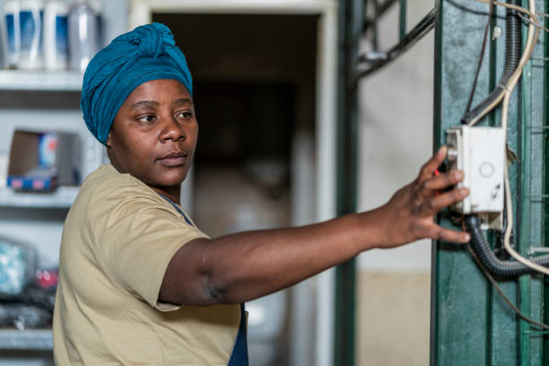 African female worker with traditional headscarf in a factory, using machinery control panel inside the working space, pushing a button.