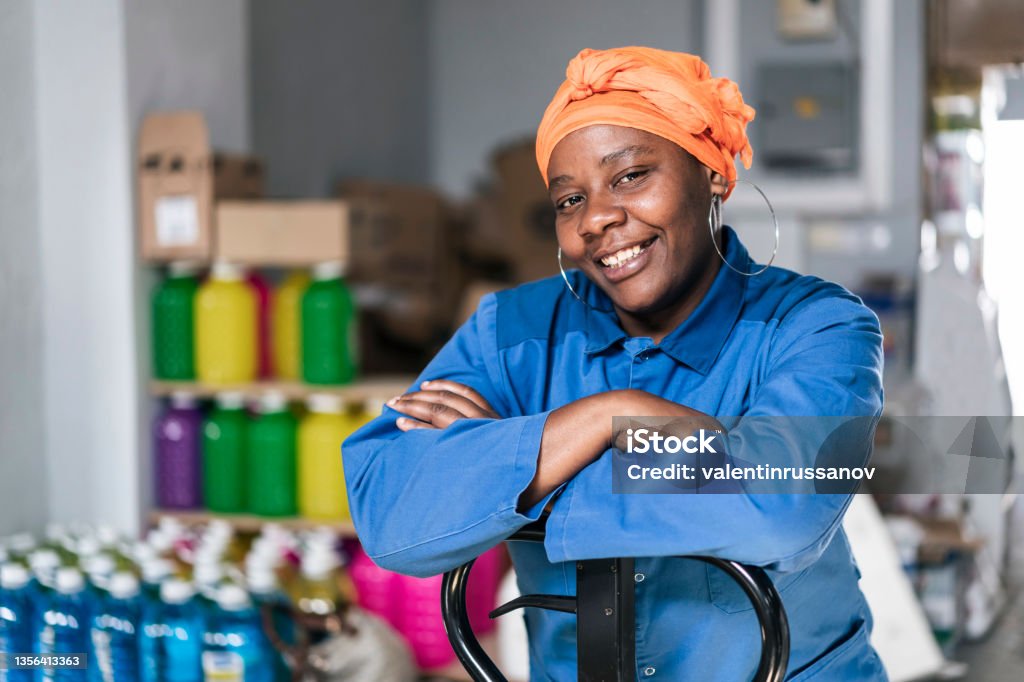 Mid adult african woman with hand pallet standing in a distribution warehouse. Portrait of a happy afro woman with headscarf, wearing uniform, working in a factory warehouse. African Ethnicity Stock Photo