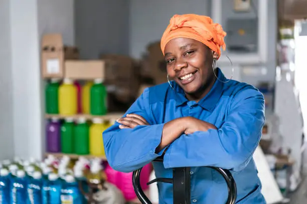 Mid adult african woman with hand pallet standing in a distribution warehouse. Portrait of a happy afro woman with headscarf, wearing uniform, working in a factory warehouse.