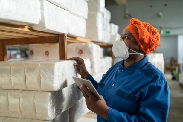 istock Female warehouse worker wearing protective face mask, holding digital tablet. African american woman in uniform working in large distribution warehouse. 1356413364