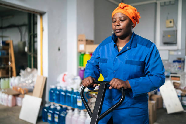 istock Female african worker with working uniform and traditional headscarf,  pulling hand truck with boxes through  a warehouse 1356413372