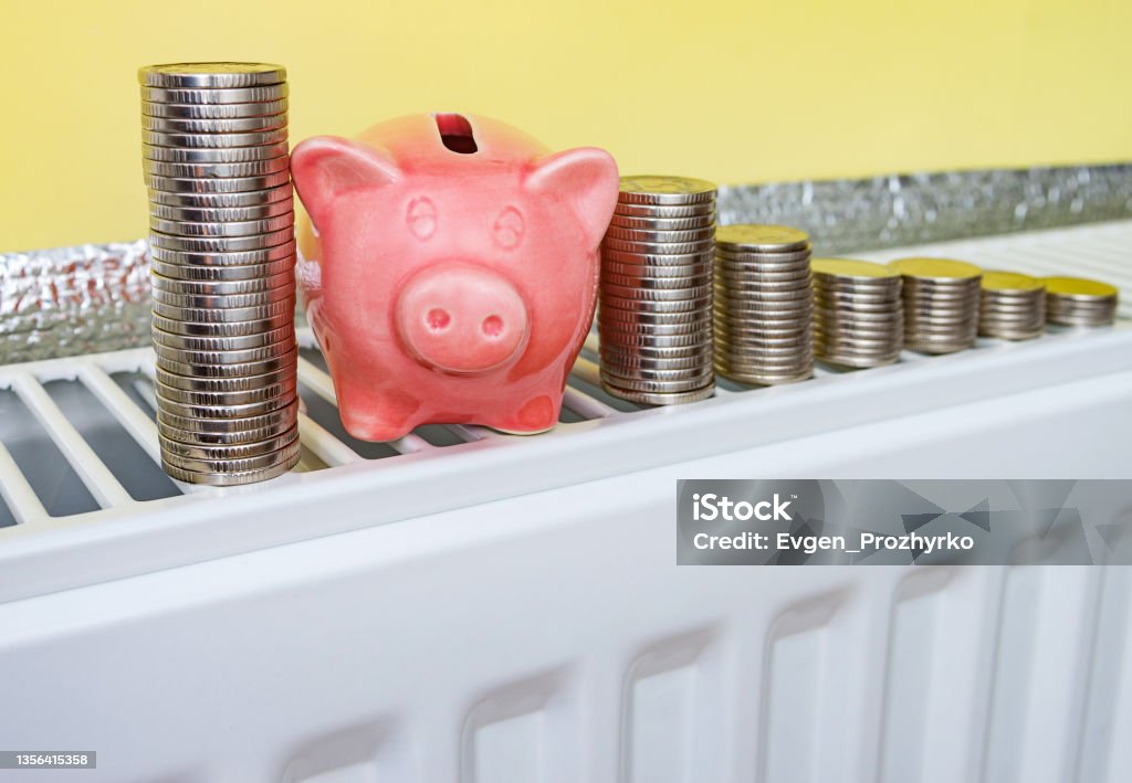 Pink piggy bank with coins on the heating radiator at home. Saving family budget, natural gas and money for heating at winter cold time. Concept image. Public Utility Stock Photo