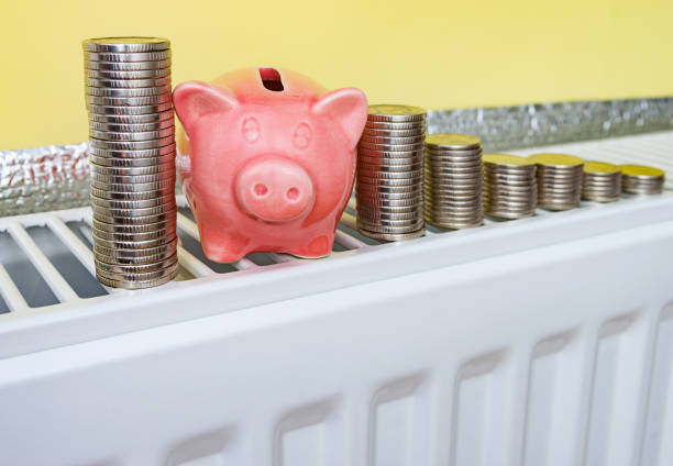 Pink piggy bank with coins on the heating radiator at home. Saving family budget, natural gas and money for heating at winter cold time. Concept image. public utility stock pictures, royalty-free photos & images