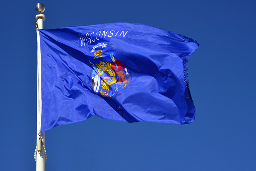 Milwaukee, Wisconsin, USA: Wisconsin state flag fluttering in the wind against blue sky (real photo, not computer generated) - Shield on a dark blue field with the state motto \
