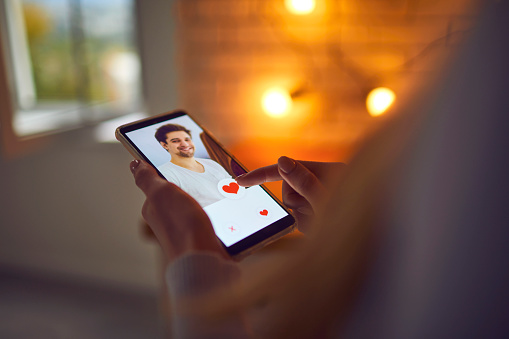 Looking for perfect guy. Woman pressing red heart like button below handsome young man's profile photo on dating app. Close-up of female hands holding phone. Find love online concept. Blur, soft focus