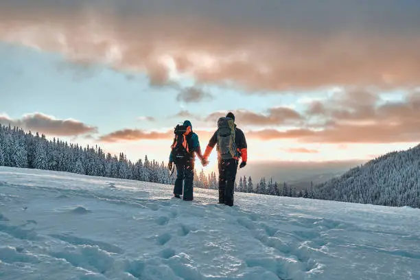 Photo of Rest in the mountains. Family couple holding hands and walking in the snowy pine mountains at sunset. The concept of recreation and tourism in winter.