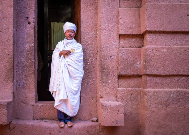 Two boys waiting outside a church, Lalibela, Ethiopia Lalibela, Ethiopia - May 23, 2021: One priest outside a church, holding a golden cross and wearing white clothing and a turban ethiopian orthodox church stock pictures, royalty-free photos & images