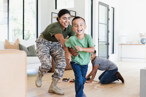 While the preteen boy sets up the wooden train set on the living room floor, the female soldier chases the elementary age boy through the house.  Everybody is smiling and happy.