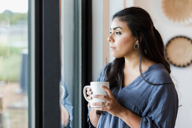 mid adult woman stands at window with hot drink - depression women sadness window imagens e fotografias de stock