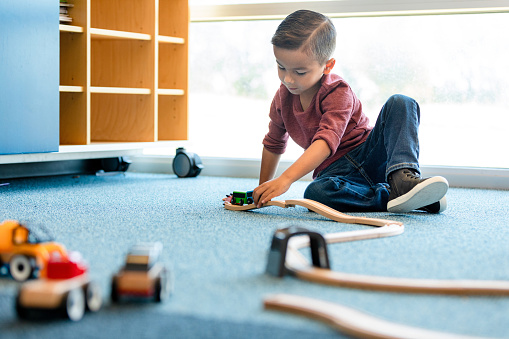 The preschool age boy contentedly plays with the toy trains on the floor of his classroom.