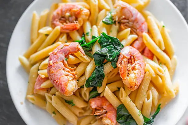 Macro closeup flat lay top view of sauteed cooked whole large king jumbo Argentinian shrimp seafood with penne Italian pasta in olive oil sliced garlic with green basil herb