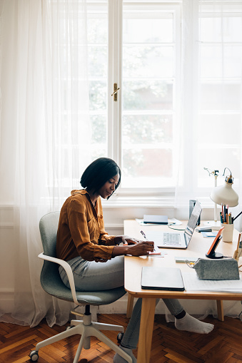 Serious African-American businesswoman writing notes in a notebook while sitting at desk with laptop computer in a living room.