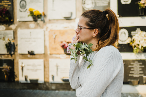 Side view of pensive adult female with ponytail in casual clothes and sunglasses touching lips and looking away sadly while standing near gray wall with bunch of flowers