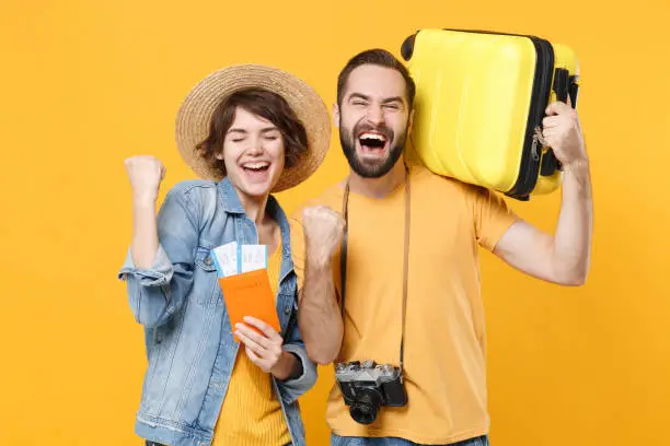 Photo of Happy young tourists couple friends guy girl isolated on yellow background. Passenger traveling abroad on weekends. Air flight journey concept. Hold passport tickets suitcase doing winner gesture.