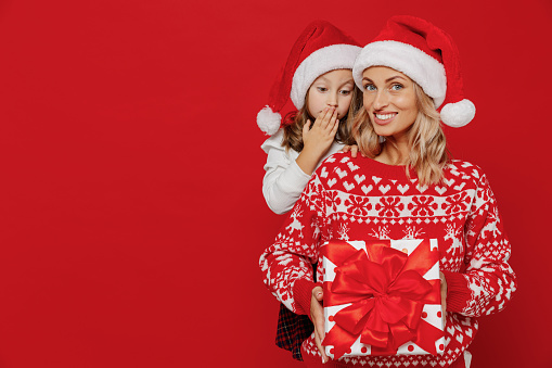Young amazed woman in sweater hat have fun with child baby girl 6-7 years old. Mommy little kid daughter hold red gift present box isolated on plain red background studio. New Year love family concept