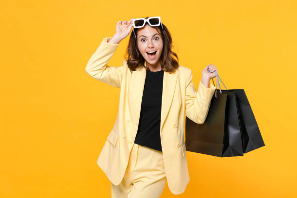 istock Excited young brunette woman in basic light suit jacket eyeglasses hold package bags with purchases after shopping keeping mouth open isolated on yellow background studio portrait. Black friday sale. 1356445119