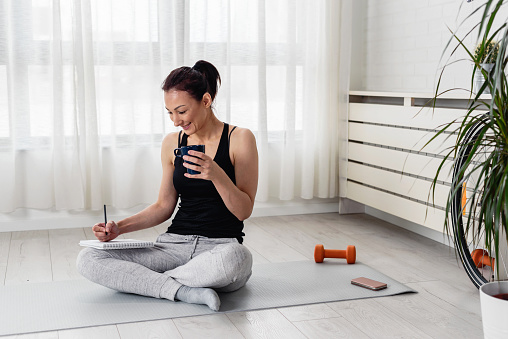 Young woman writing in her notebook. Sitting at home by the window on a yoga mat after exercise. Personal growth resolutions