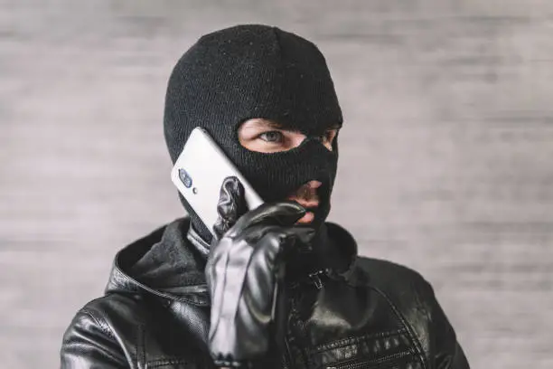 The thief is talking on the phone in a black mask. The extortionist demands money for the kidnapping. a phone fraudster commits a crime, violates the law. concept of telephone terrorism and fraud.