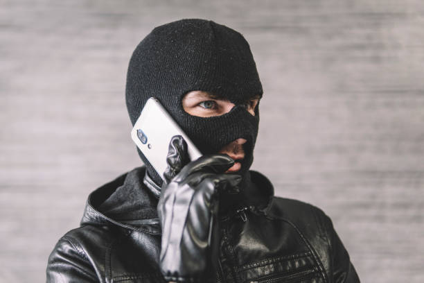 thief is talking on the phone in a black mask. the extortionist demands money for the kidnapping. a phone fraudster commits a crime, violates the law. concept of telephone terrorism and fraud. - extortionist imagens e fotografias de stock