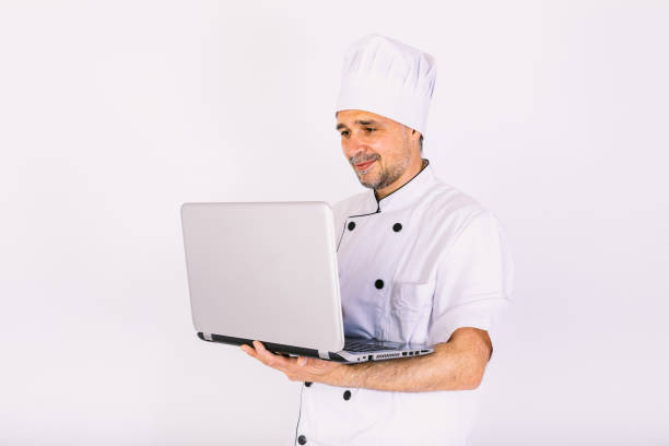 istock Chef cook wearing kitchen jacket and hat, holding a laptop and showing its screen, on white background 1356462123