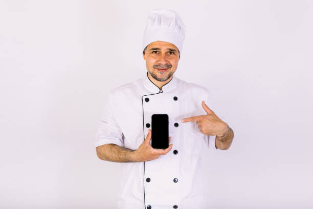 istock Chef dressed in white hat and jacket showing his mobile phone on white background. Restaurant, food and takeaway concept. 1356462281