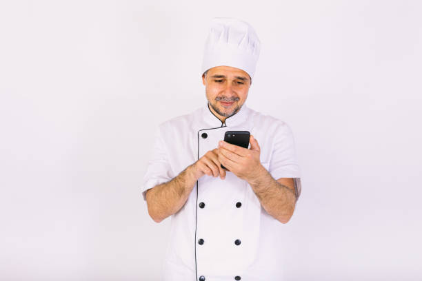 istock Cook dressed in white hat and jacket looking at his mobile phone on white background. Restaurant, food and takeaway concept. 1356462290
