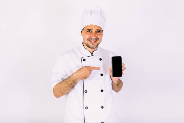 istock Chef dressed in white hat and jacket showing his mobile phone on white background. Restaurant, food and takeaway concept. 1356462347