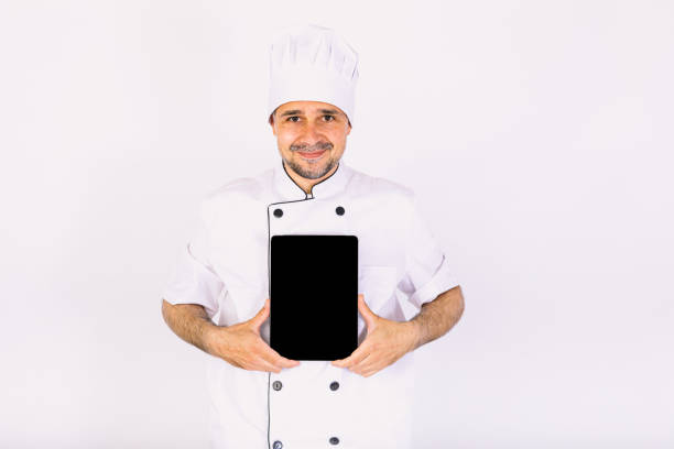 istock Chef cook wearing kitchen jacket and hat, holding a tablet, showing its screen, on white background 1356462372