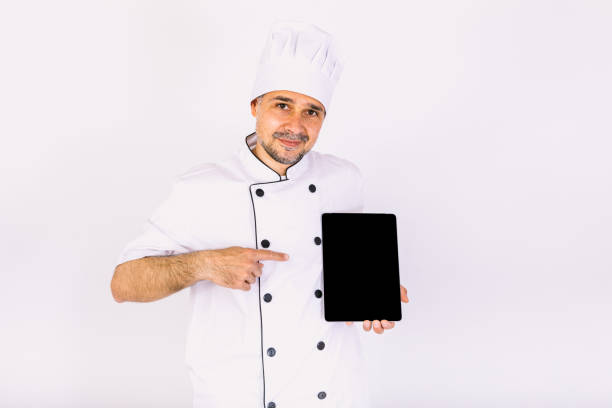 istock Chef cook wearing kitchen jacket and hat, holding a tablet, showing its screen, on white background 1356462391