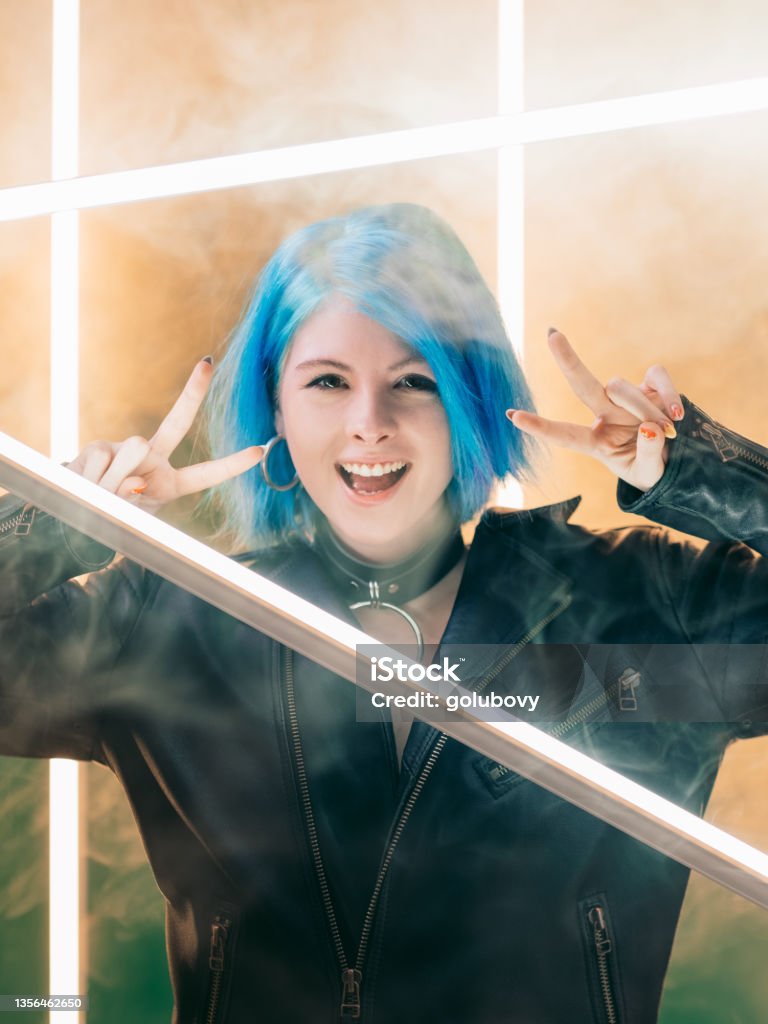peace gesture cyberpunk party woman led light Peace gesture. Cyberpunk party. Synthwave style. Excited smiling woman showing victory hand sign on LED lamp light on smoke background. Beauty Stock Photo
